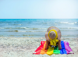 a woman sitting on her back on the seashore on a pride flag. she is wearing a straw hat and yellow scarf. lgtb. concept of diversity and tolerance.