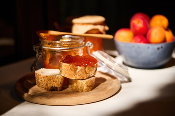 Daylight falls on part of the table with whole grain bread with apricot jam and ripe fruits in a bowl. Rustic still life
