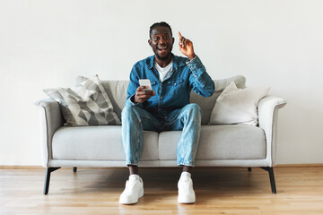 Excited African American Guy Holding Smartphone Having Idea Sitting Indoors