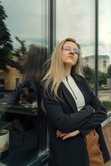 A beautiful woman in glasses stands leaning against the window of a building, looking into the camera.
