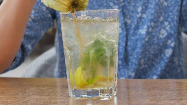 Summer cocktail. Mojito cocktail in a transparent glass glass with pieces of ice, a mint and lemon leaf, a woman drinks a cooling cocktail from a cocktail tube on the veranda of a summer cafe