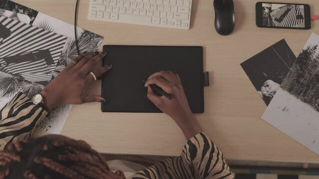 Top view slowmo of unrecognizable African American female photographer drawing on graphic tab sitting at office desk with printed black and white photos on it