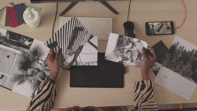 Top view slowmo of unrecognizable African American female retoucher looking at beautiful printed black and white photos while working at office desk in front of computer
