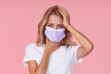 Young ill woman in a white casual t-shirt wears a protective face mask. holding hands on neck. Protection against flu and cold diseases during a pandemic. Pink isolated background