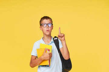 Portrait of a schoolboy in glasses with textbooks and a backpack on a yellow background showing...