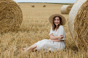 Naklejka na ściany i meble A young woman in a dress and hat posing in a field near hay bales. A woman near a haystack in a field. Summer photoset