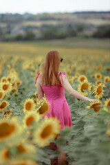 A long-haired girl in a pink dress walks through a sunflower field. The concept of vegetable oil or good harvest of sunflower seeds. The girl straightens her brown hair in windy weather.
