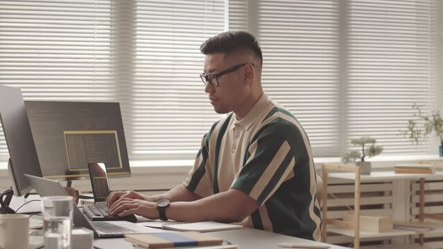 Medium slowmo portrait of male Asian developer typing program code on computer then looking at camera, sitting at office desk