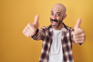 Hispanic man with beard standing over yellow background approving doing positive gesture with hand, thumbs up smiling and happy for success. winner gesture.