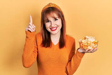 Redhead young woman holding potato chips smiling with an idea or question pointing finger with happy face, number one