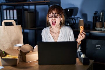 Young beautiful woman working using computer laptop and eating delivery food angry and mad...