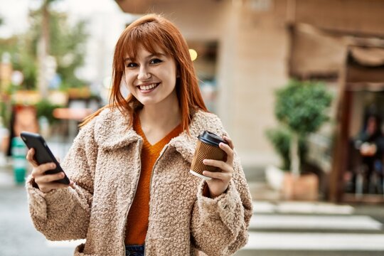 Young redhead girl smiling using smartphone drinking coffee at the city.