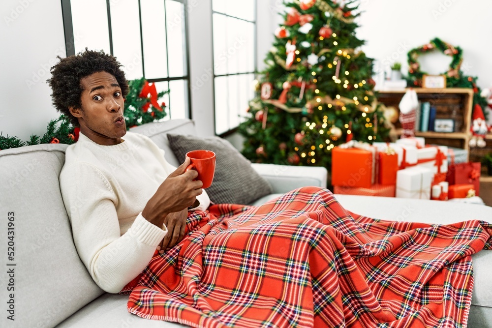 Poster Young african american man sitting on the sofa drinking coffee by christmas tree making fish face with lips, crazy and comical gesture. funny expression.