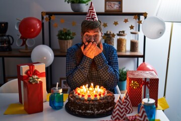 Caucasian man with long beard celebrating birthday holding big chocolate cake shocked covering mouth with hands for mistake. secret concept.