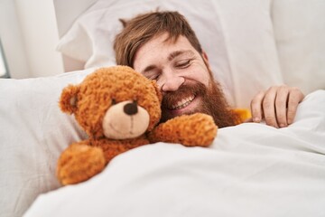 Young redhead man lying on bed hugging teddy bear at bedroom