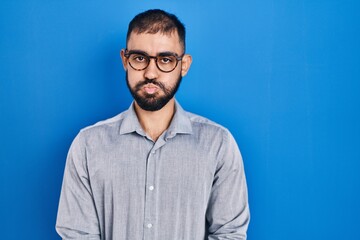 Obraz premium Middle east man with beard standing over blue background puffing cheeks with funny face. mouth inflated with air, crazy expression.