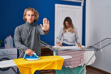 Couple ironing clothes at laundry room with open hand doing stop sign with serious and confident expression, defense gesture