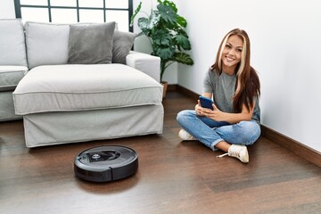 Obraz premium Young caucasian girl controlling robot vacuum cleaner with smartphone sitting on the floor at home.