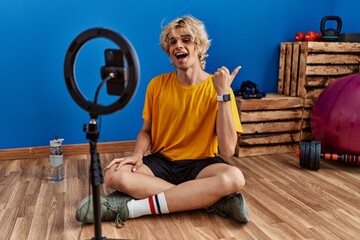 Young man recording training tutorial at the gym pointing thumb up to the side smiling happy with...