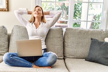 Young blonde woman relaxed with hands on head using laptop at home.