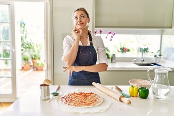 Beautiful blonde woman wearing apron cooking pizza serious face thinking about question with hand...
