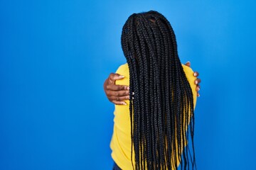 Beautiful black woman standing over blue background hugging oneself happy and positive from backwards. self love and self care