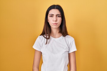 Young beautiful woman standing over yellow background puffing cheeks with funny face. mouth...