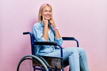 Beautiful blonde woman sitting on wheelchair smiling with open mouth, fingers pointing and forcing cheerful smile