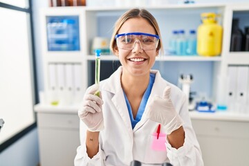 Young blonde woman working at scientist laboratory holding sample smiling happy and positive, thumb up doing excellent and approval sign