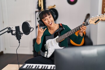 Young beautiful hispanic woman musician playing electrical guitar doing rock symbol with fingers at music studio