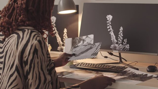 Medium slowmo of African American female retoucher in stylish zebra print shirt looking at printed black and white photos sitting by computer at her workplace