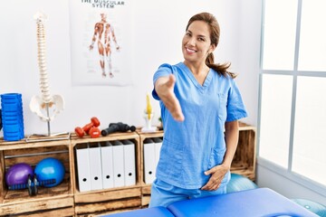 Obraz premium Middle age hispanic physiotherapist woman working at pain recovery clinic smiling friendly offering handshake as greeting and welcoming. successful business.