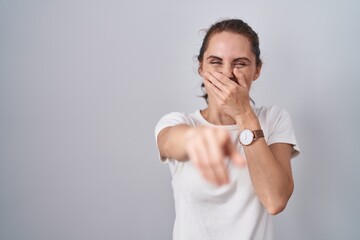 Beautiful brunette woman standing over isolated background laughing at you, pointing finger to the camera with hand over mouth, shame expression