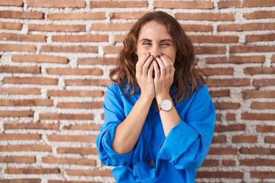 Beautiful brunette woman standing over bricks wall laughing and embarrassed giggle covering mouth with hands, gossip and scandal concept