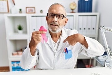 Mature doctor man holding pink cancer ribbon at the clinic pointing finger to one self smiling happy and proud