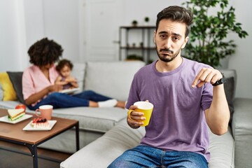 Hispanic father of interracial family drinking a cup coffee pointing down looking sad and upset, indicating direction with fingers, unhappy and depressed.