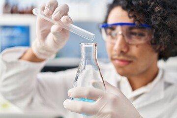 Young hispanic man wearing scientist uniform pouring liquid on test tube at laboratory