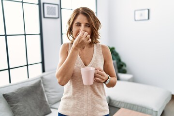 Middle age woman drinking a cup coffee at home smelling something stinky and disgusting,...