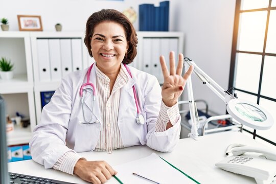 Middle age hispanic woman wearing doctor uniform and stethoscope at the clinic showing and pointing up with fingers number four while smiling confident and happy.