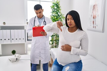 Young hispanic mother expecting a baby at the doctor with angry face, negative sign showing dislike with thumbs down, rejection concept