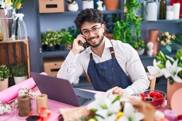 Young hispanic man florist using laptop talking on smartphone at florist shop