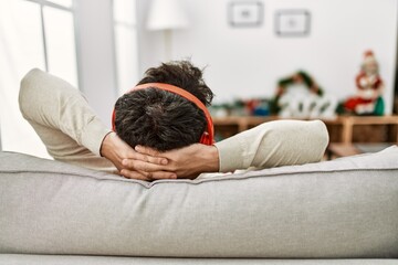 Young hispanic man on back view using headphones sitting on the sofa with hands on head at home.