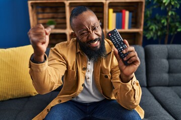 African american man holding television remote control annoyed and frustrated shouting with anger,...