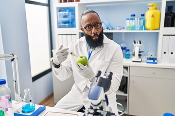 African american man working at scientist laboratory with apple making fish face with mouth and...