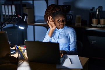 African woman working at the office at night smiling with hand over ear listening an hearing to rumor or gossip. deafness concept.