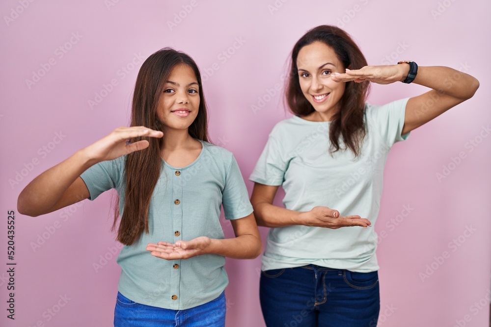 Sticker young mother and daughter standing over pink background gesturing with hands showing big and large s