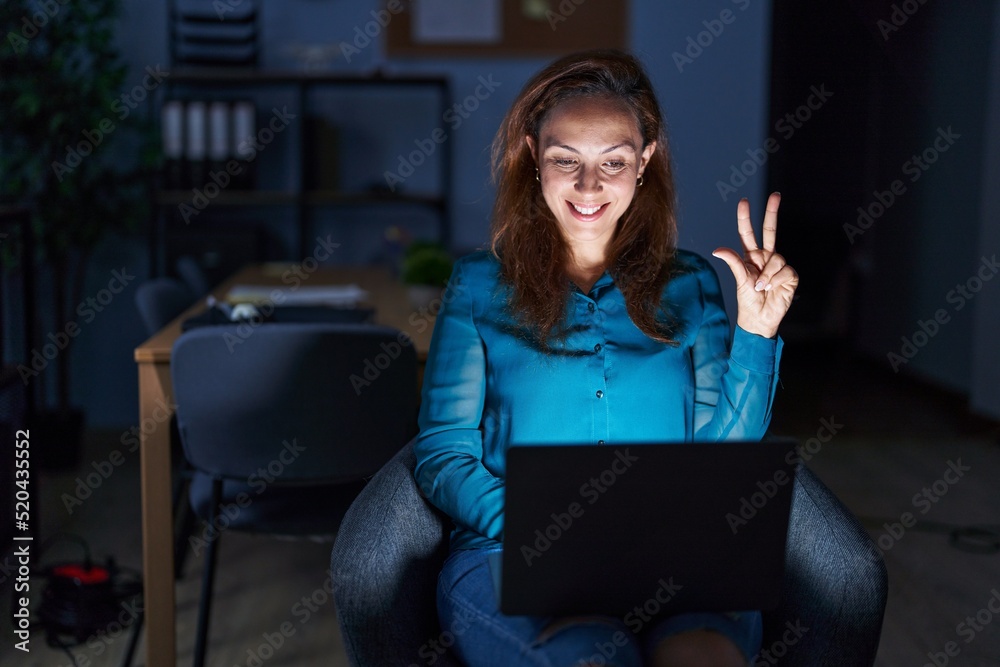 Wall mural Brunette woman working at the office at night showing and pointing up with fingers number three while smiling confident and happy.