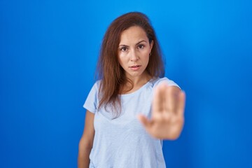 Fototapeta premium Brunette woman standing over blue background doing stop sing with palm of the hand. warning expression with negative and serious gesture on the face.