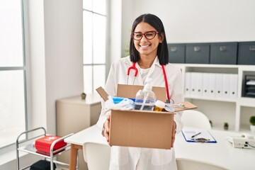 Young hispanic doctor woman holding box with medical items winking looking at the camera with sexy...
