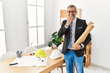 Middle age hispanic business man holding paper blueprints laughing and embarrassed giggle covering...